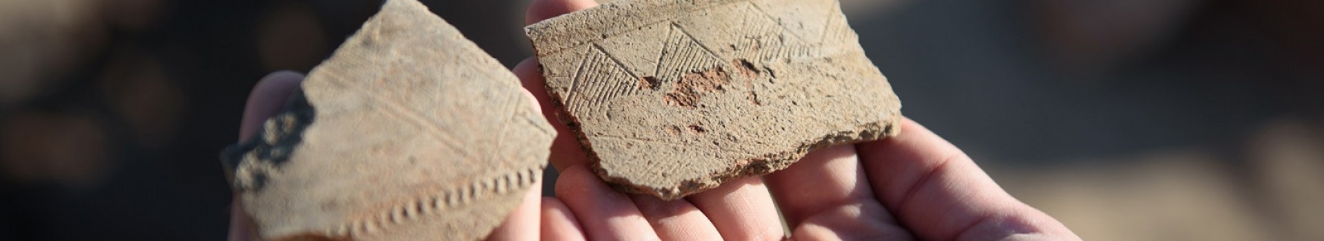 Two hands hold fragments of recently excavation pottery with a zig-zag design.