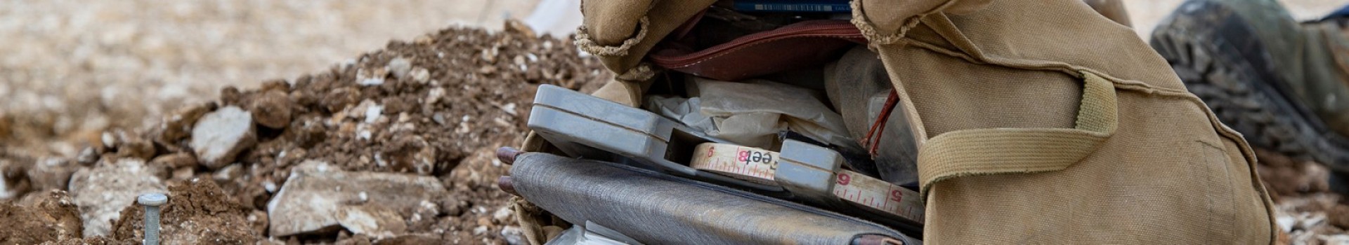 An archaeologist's bag containing a tape measure lies on the earth by a booted foot.