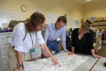 Member-level archaeologists examine plans in an office.