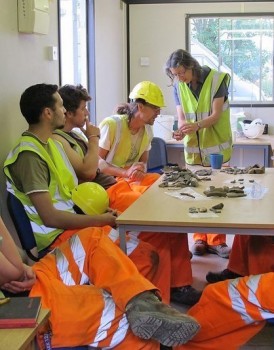 Archaeologists in high-visibility clothing sits around a table and examine finds.